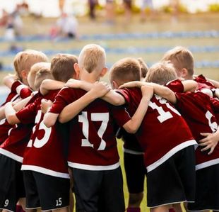 A team of children in matching maroon jerseys with white numbers huddled together in a circle on a sports field.