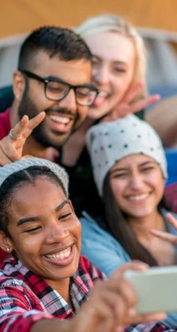 A group of four friends smiles while taking a selfie together, making peace signs with their hands.