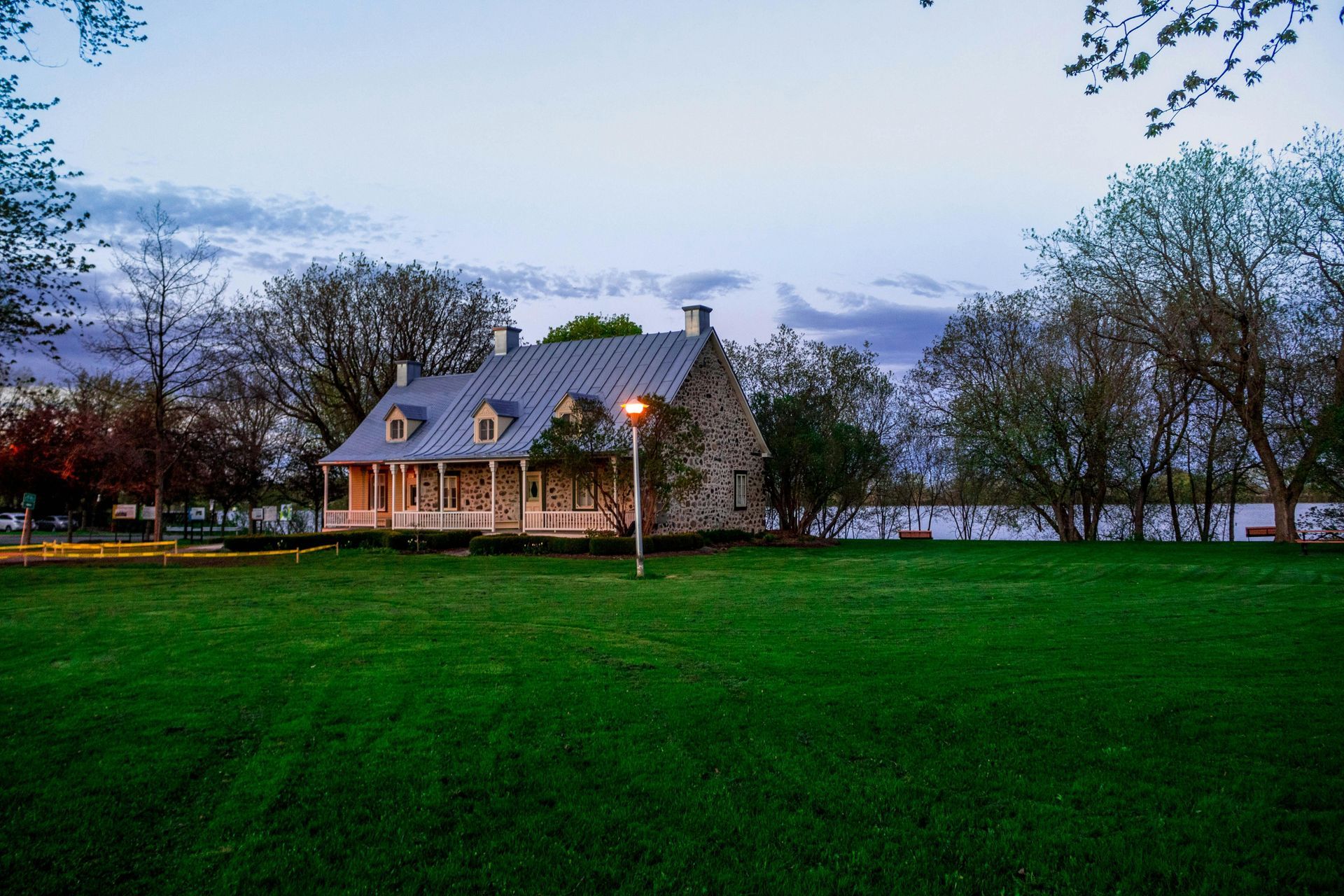 Stone house with dormers, set in a green lawn, surrounded by trees. Dusk sky over a lake.