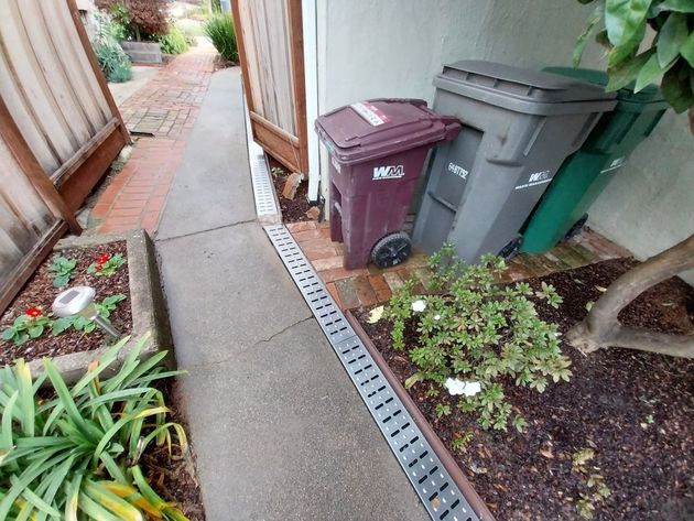 Narrow side yard walkway beside houses, with trash bins, plants, and a drainage channel.
