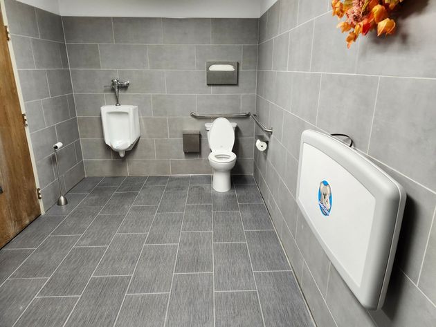 Accessible public restroom with gray tile walls, a urinal, toilet, and fold-down changing table