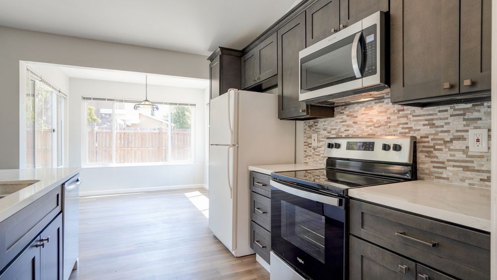 Bright modern kitchen with gray cabinets, stainless appliances, and a sunlit dining nook beyond.