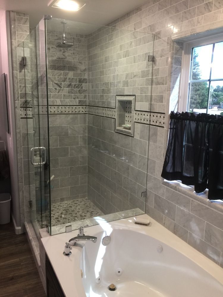 Gray tiled bathroom with glass shower, white bathtub, and window with dark curtain