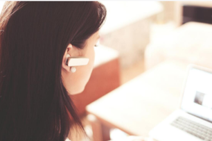 Picture of a woman provide remote computer technical support. The woman has brown hair and is wearing a white blouse, she is looking at a laptop talking into a bluetooth ear device.