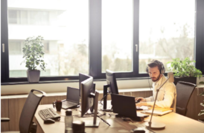 Picture of a man with a yellow shirt sitting at a desk looking at his computer. The man is wearing headphones and speaking to someone on the computer. He is sitting at a desk with multiple laptops with no one else sitting with him. The room is bright with windows surrounded with plants.