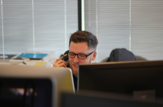 Picture of a man with glasses speaking on the phone. The man is sitting at a computer desk with multiple computer screens.