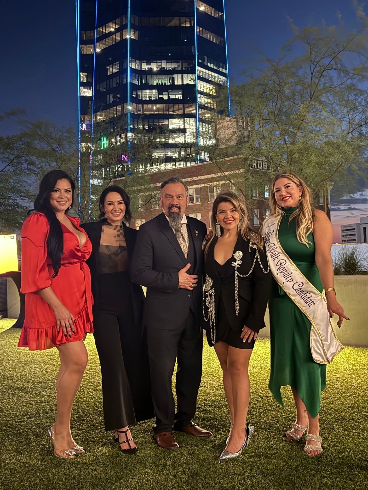 Five people in formal attire posing outdoors at night in front of a lit high-rise building.