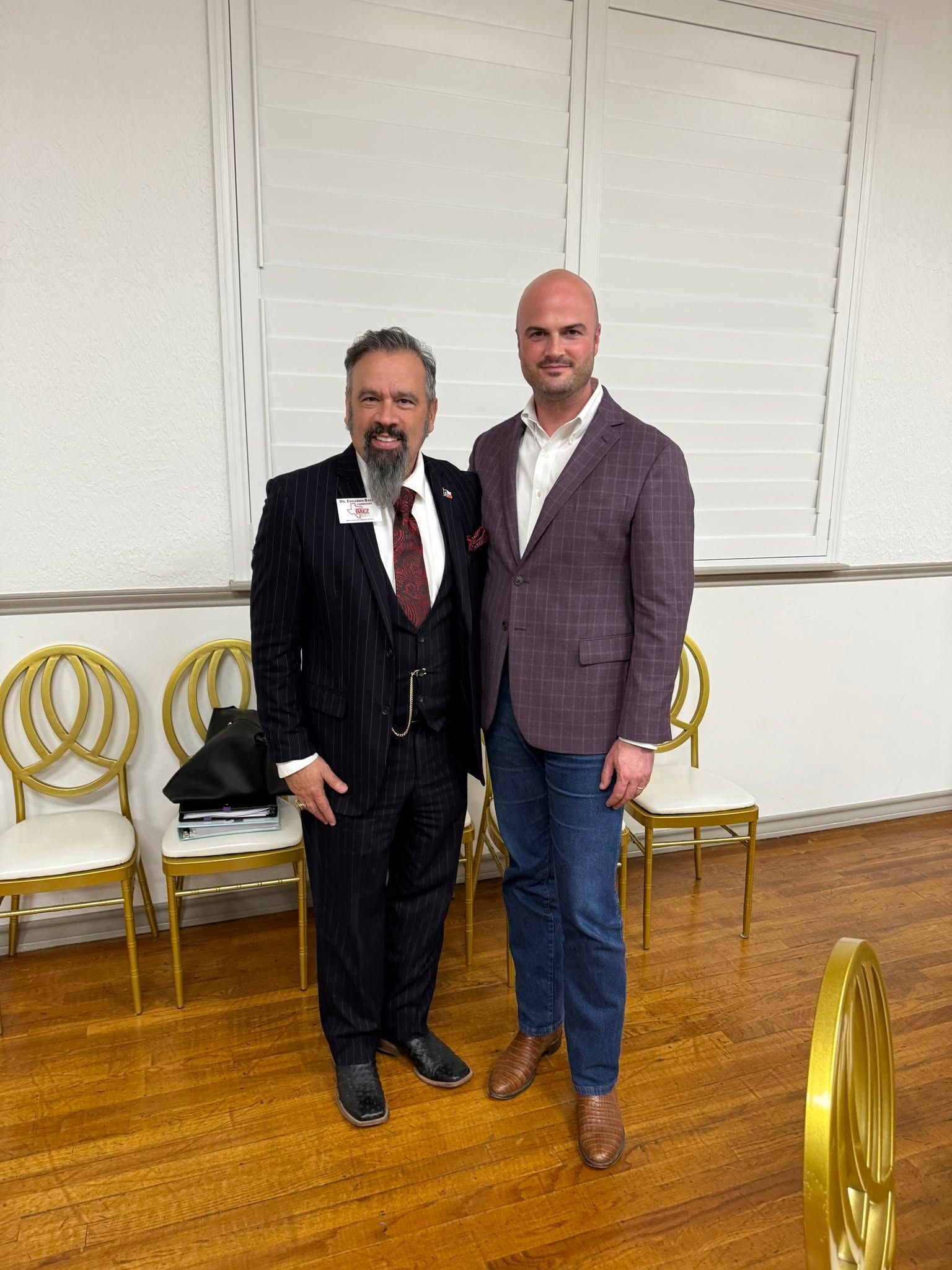 Two men posing together indoors, one in a dark suit and one in a purple blazer, with gold chairs behind them
