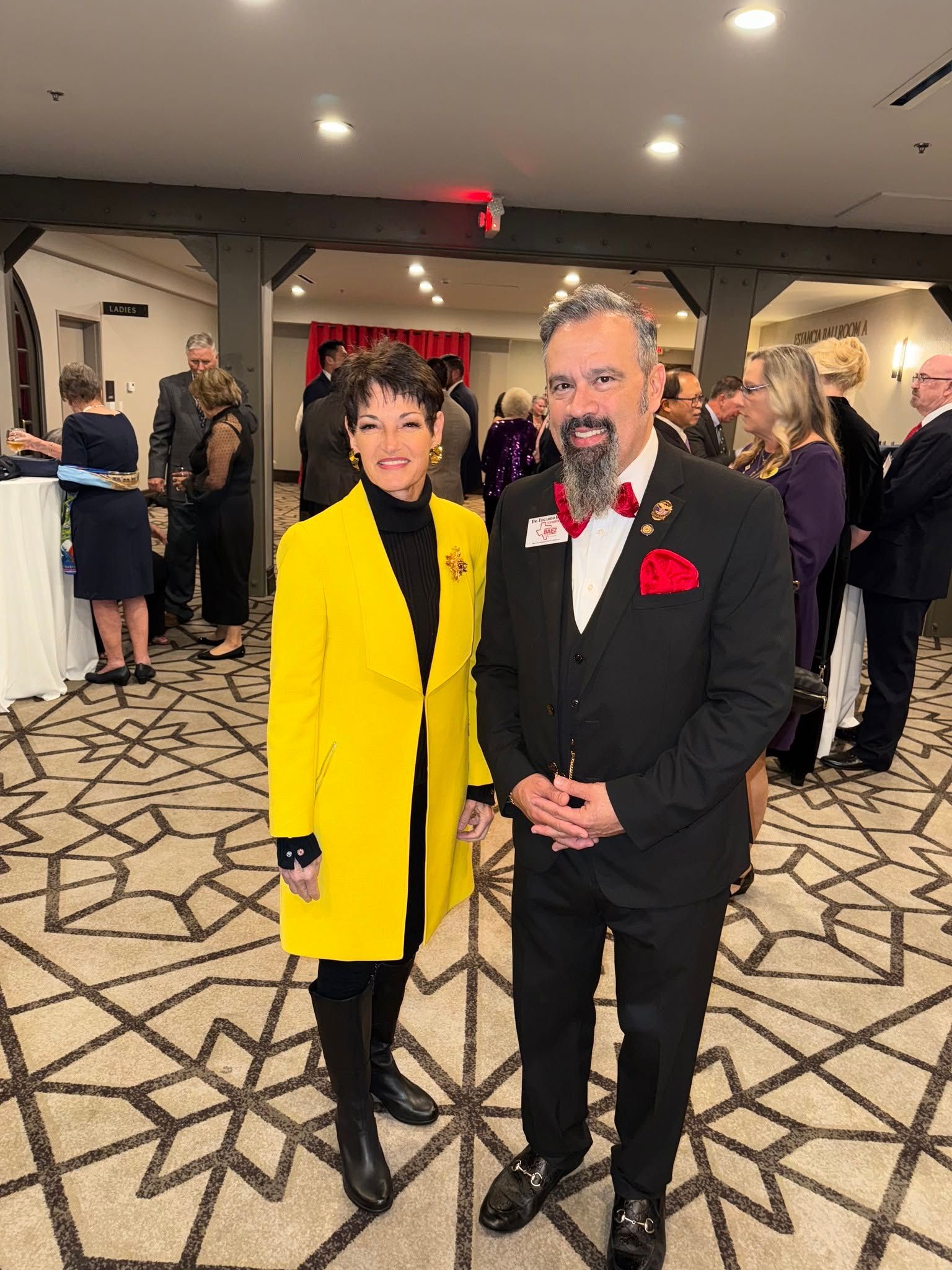 Two people in formal attire posing at an indoor event, one in a yellow coat and one in a black suit with a red boutonniere.