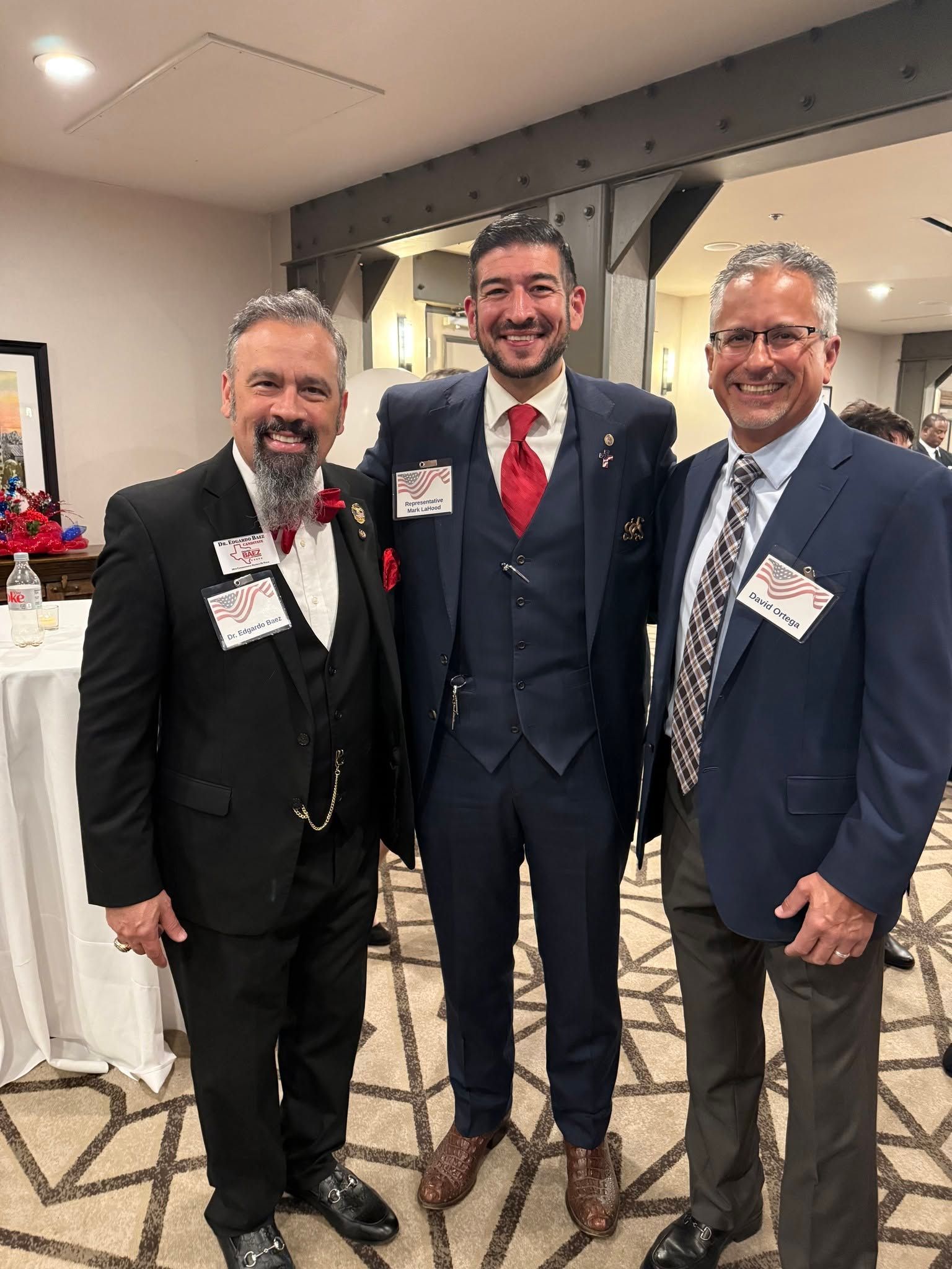 Three men in suits smiling at a formal indoor event, standing beside a decorated table.