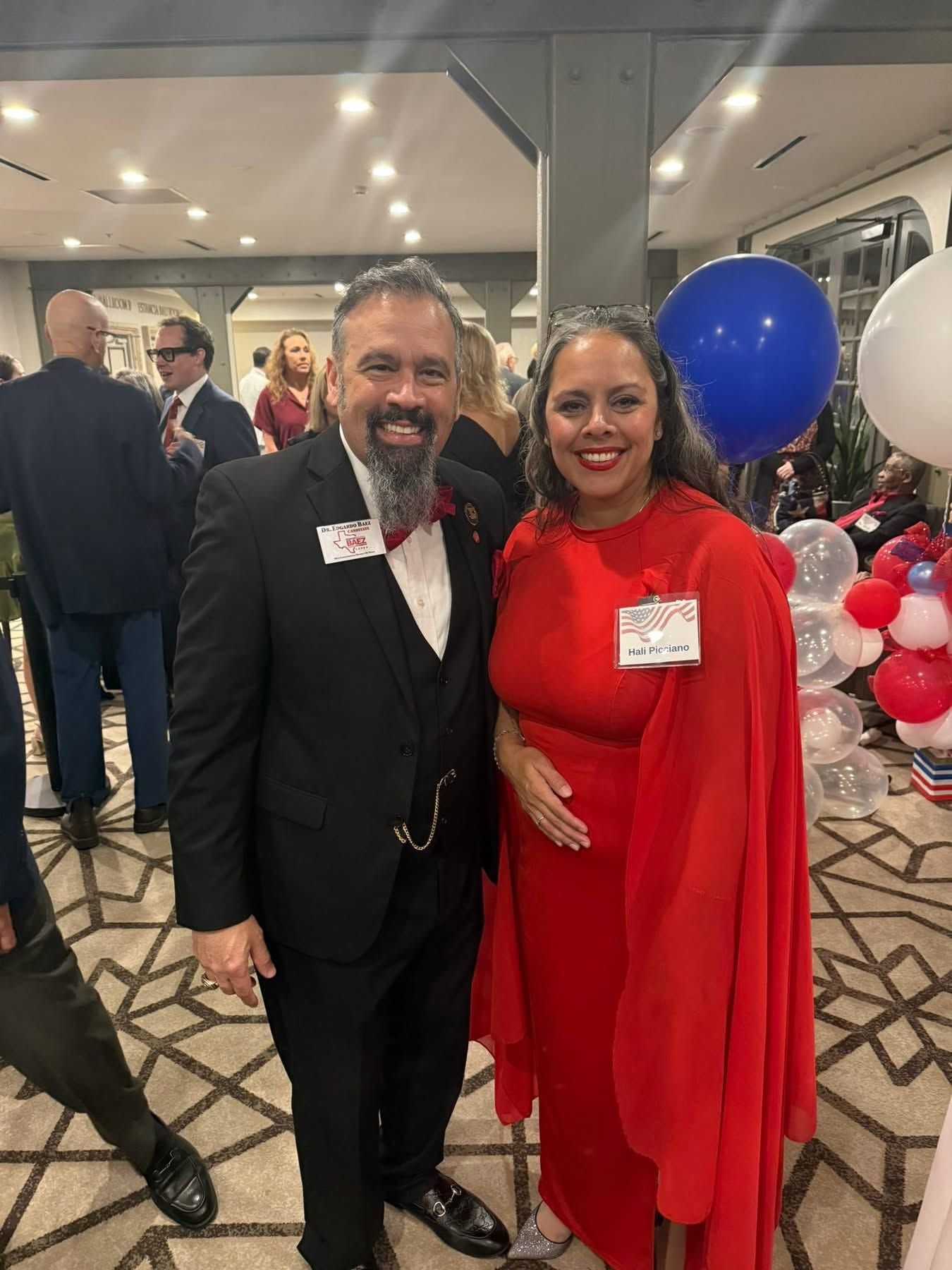 Two people dressed for a formal event, smiling indoors with red, white, and blue balloons behind them.