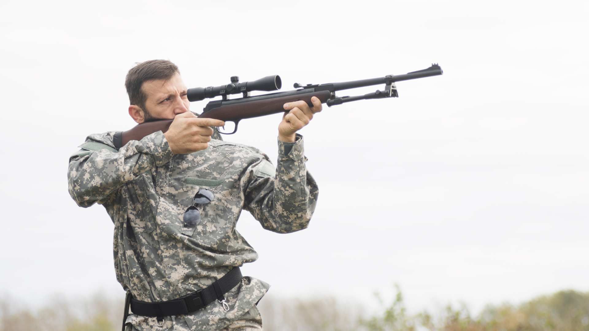 Camouflaged soldier aiming a rifle outdoors under an overcast sky