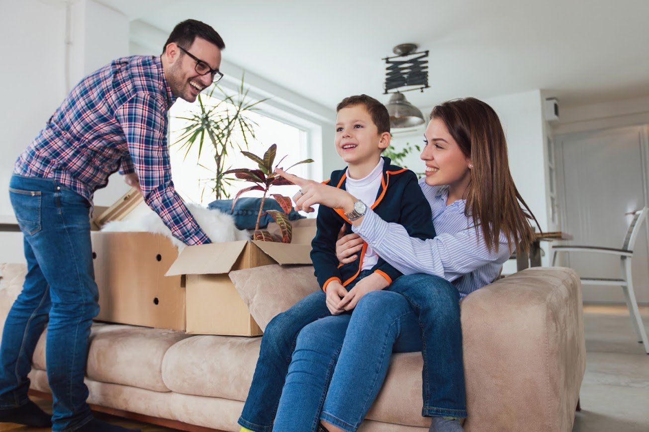 Family in new home; dad unpacking, son pointing, mom smiling on couch.