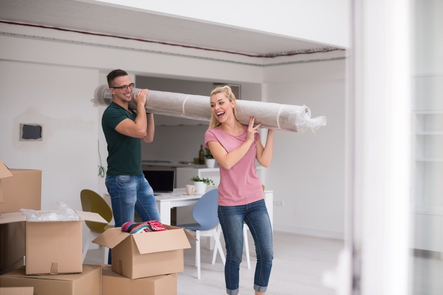 Couple carrying rolled-up carpet indoors during a move; boxes nearby, bright room.