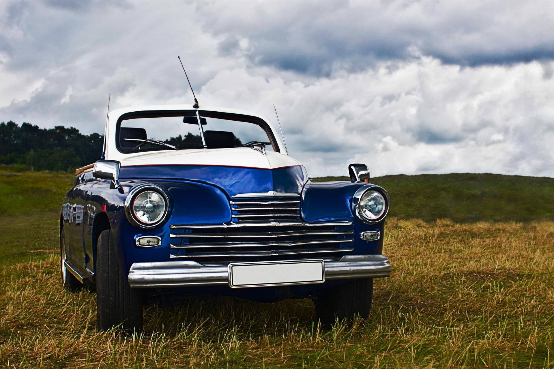 Blue and white vintage convertible car parked in a grassy field under a cloudy sky.