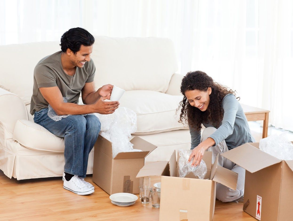 Couple unpacking boxes in a bright room, smiling as they look at contents.
