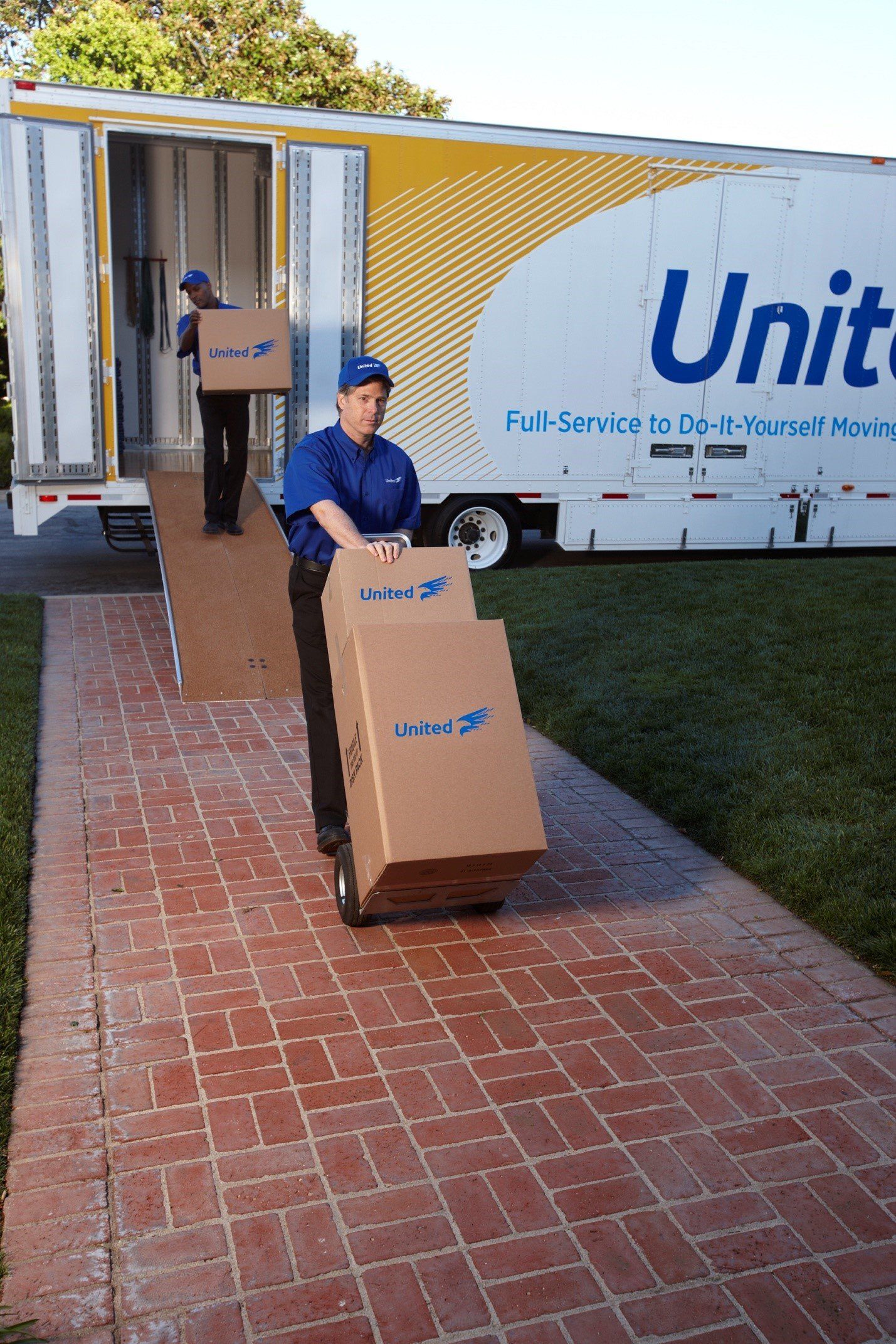 Movers in blue uniforms load boxes from a United moving truck onto a brick path.