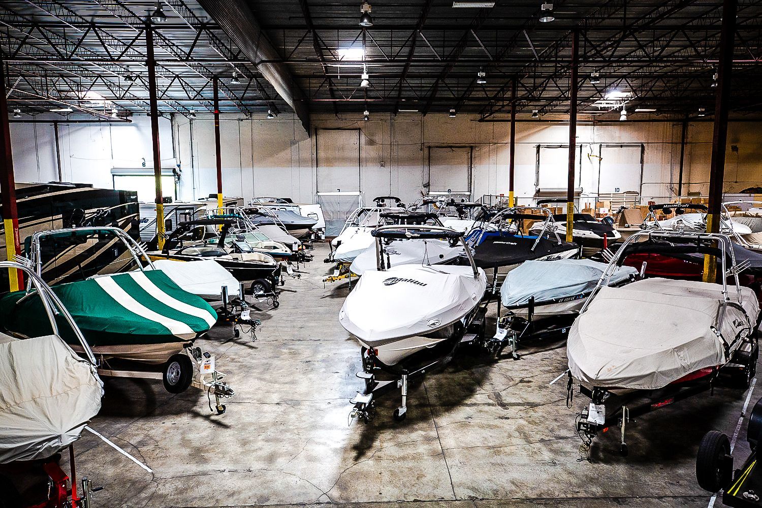 Indoor storage of boats in a large warehouse. Many boats are covered, in varying colors, and stored on trailers.