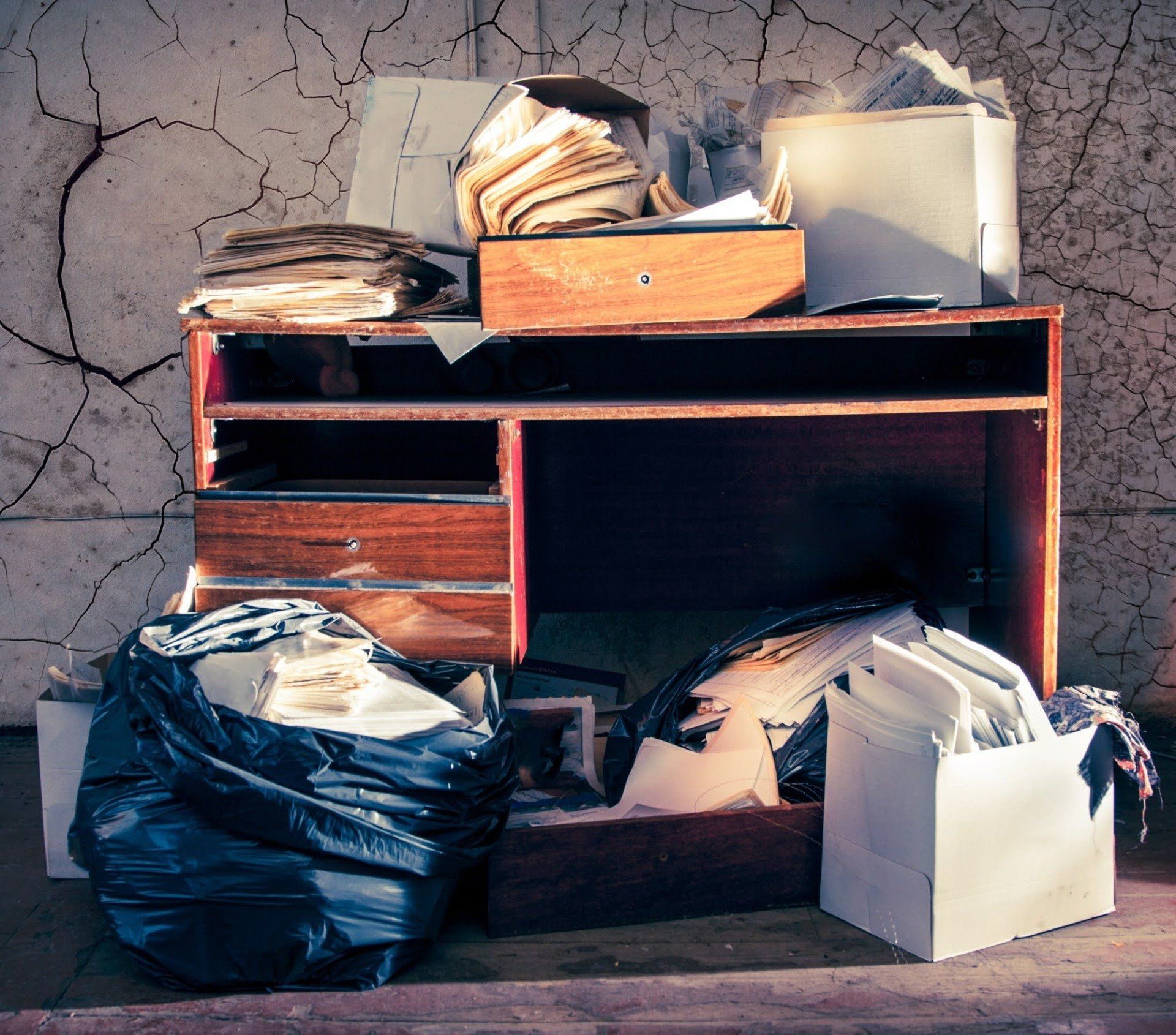 Messy desk with overflowing papers, a black trash bag, and a cracked wall.