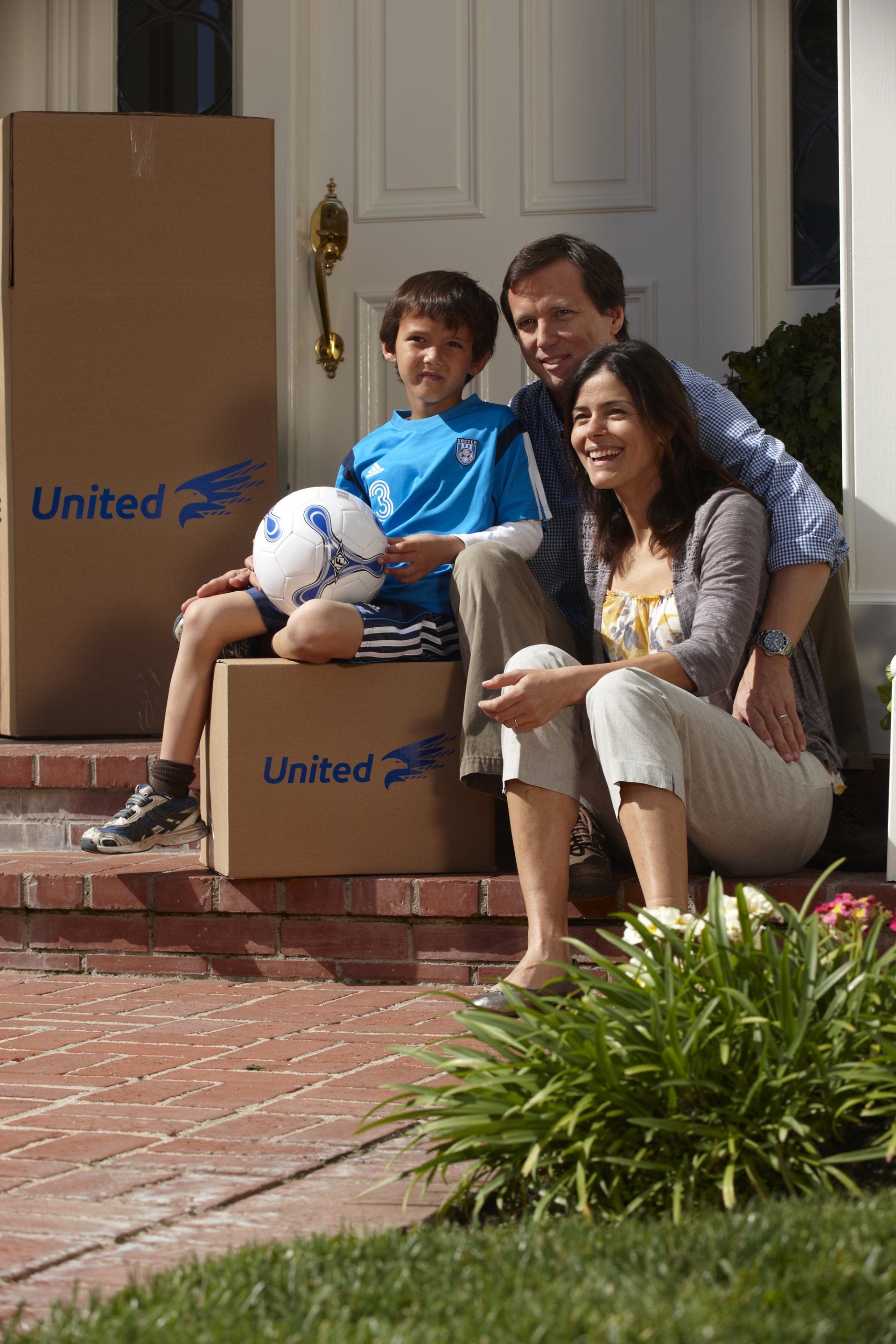 Family sitting on porch steps with moving boxes; son holds soccer ball.