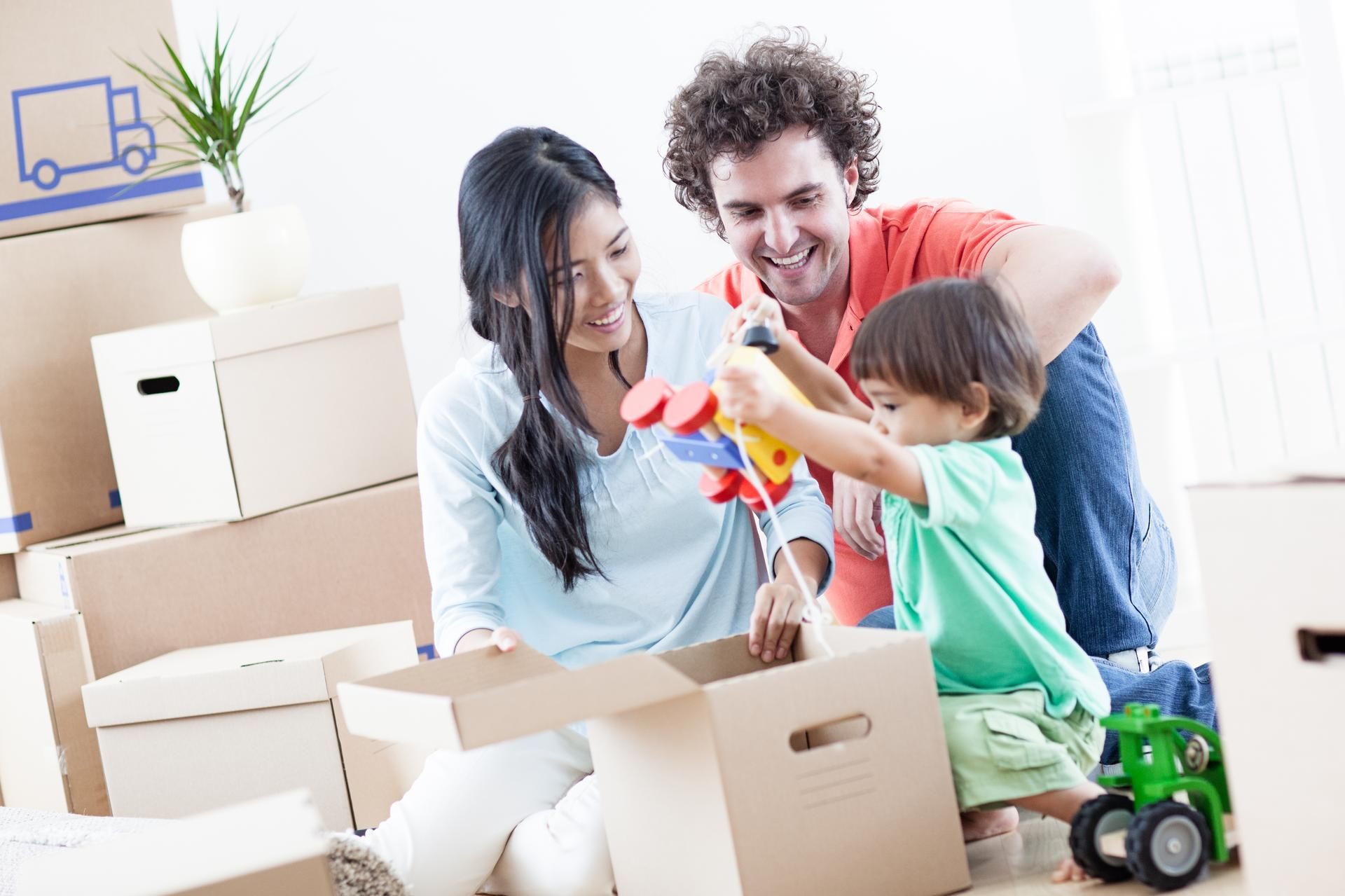 Family unpacking boxes in a new home; smiling parents watch toddler play with a toy.