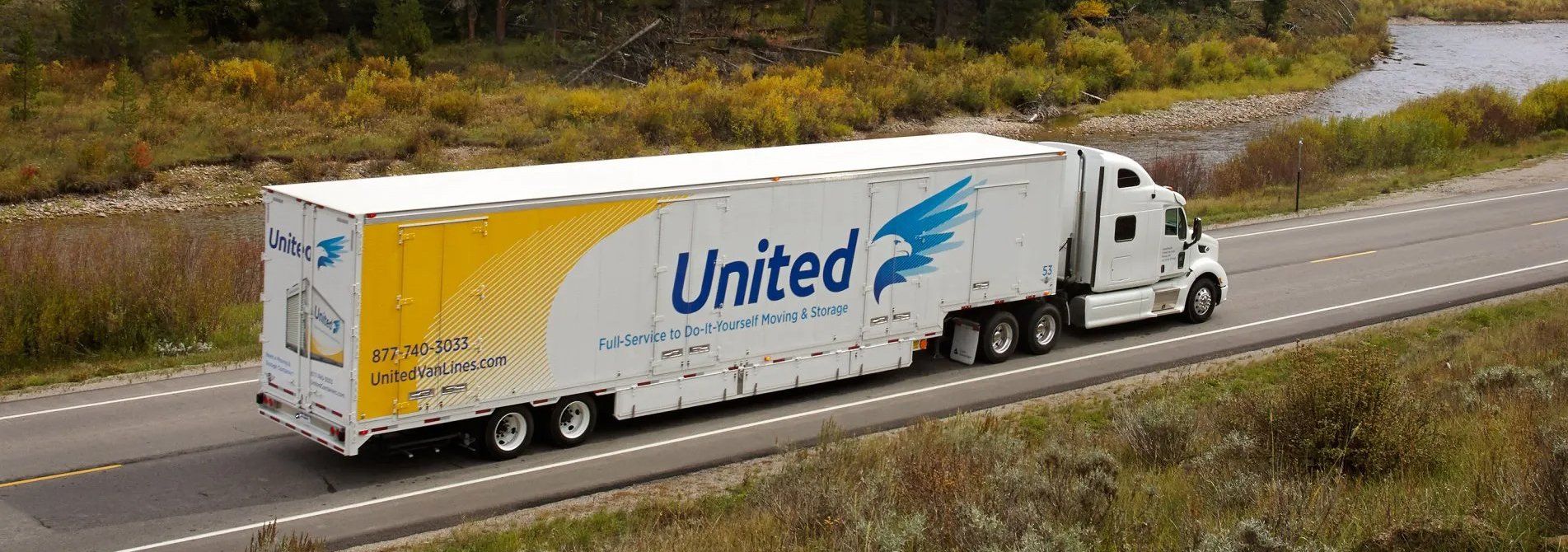 A United van lines semi-truck traveling on a road surrounded by autumn foliage.