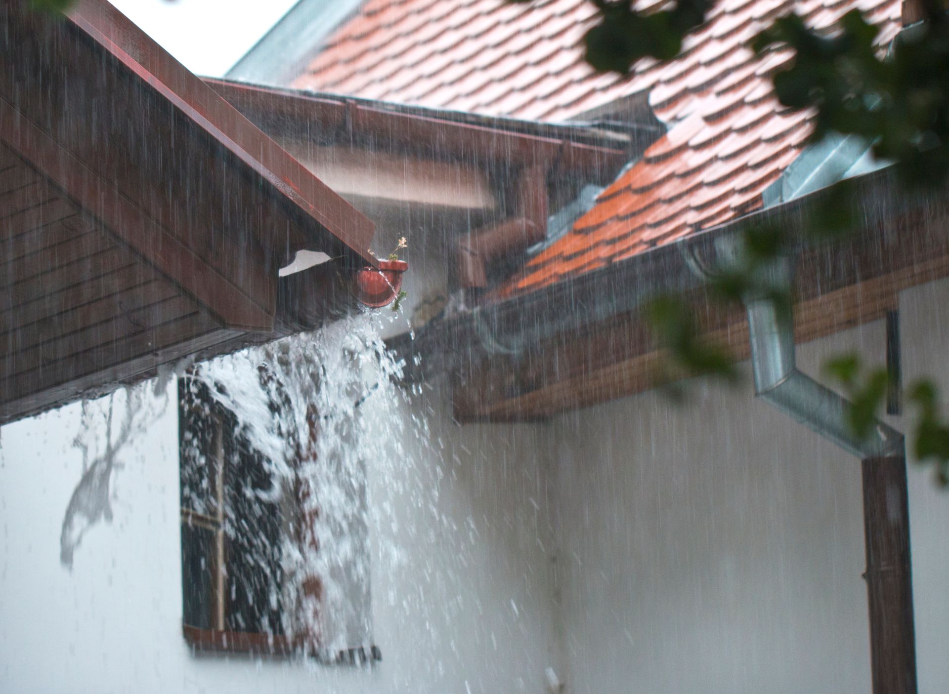Heavy rain overflowing from a clogged gutter on a white house, splashing down near a window.