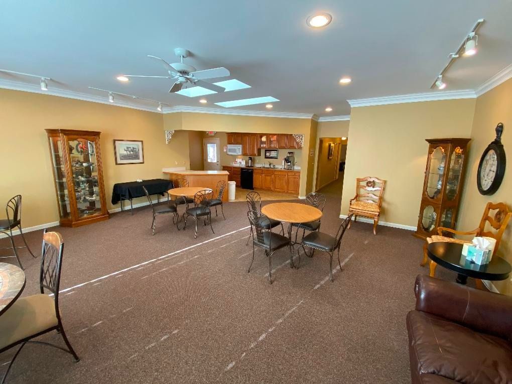 Lounge area with tables, chairs, cabinets, a small kitchen, and a pathway. Brown carpet and tan walls.