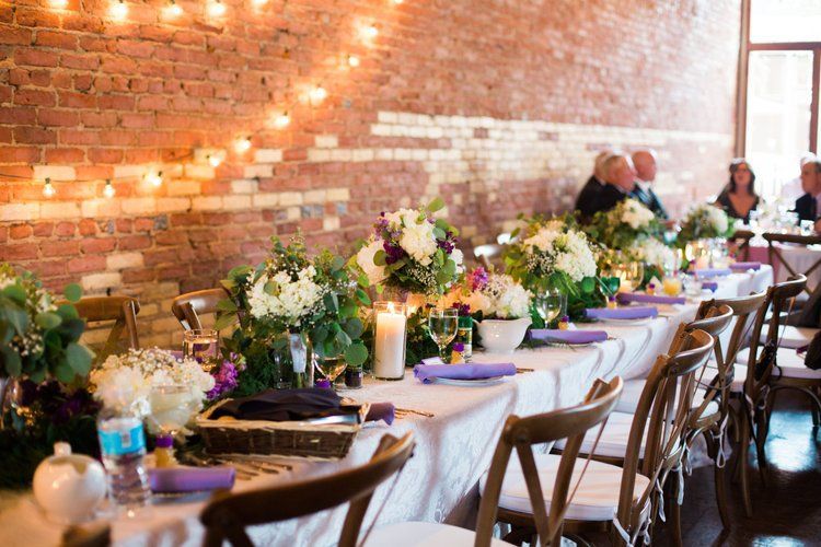 Long dining table set for a wedding with floral centerpieces, candles, and string lights against a brick wall.