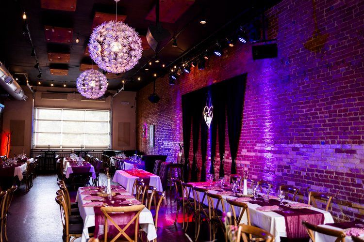 Elegant dining room with tables set for a formal event. Purple lighting against brick walls and decorative hanging lights.