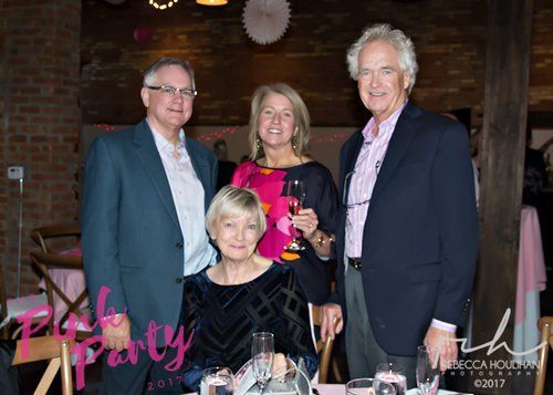 Four people at a pink-themed event, posing for a photo. A woman sits while others stand; smiling.