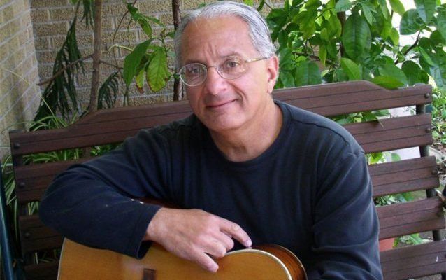 Man with glasses, guitar on lap, sitting on a wooden bench outdoors.