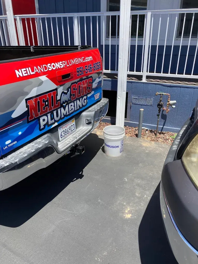A plumbing truck parked near a building, next to a bucket. The truck has a red, white, and blue logo.
