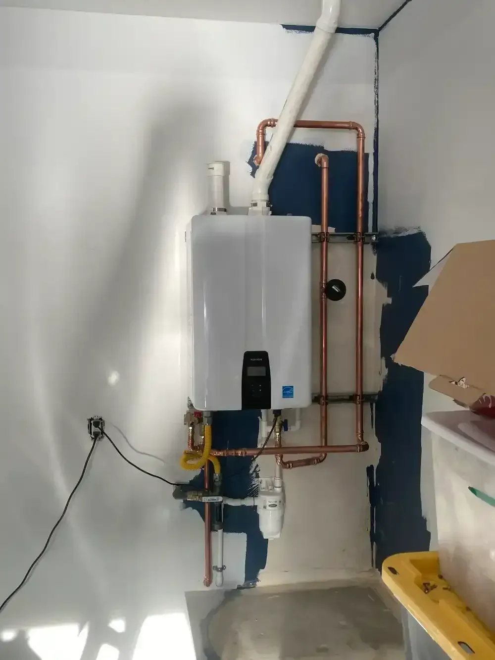 White tankless water heater mounted on a blue wall with copper pipes, a gas line, and a PVC vent pipe.