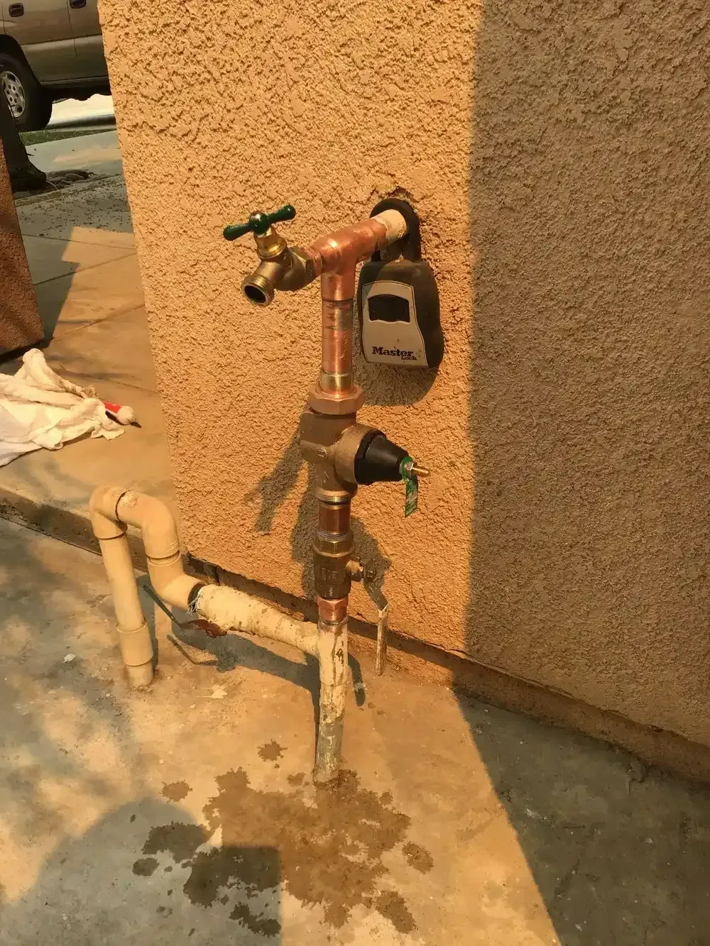 Outdoor water valve system with a copper pipe, spigot, and timer attached to a stucco wall. Water is on the ground.