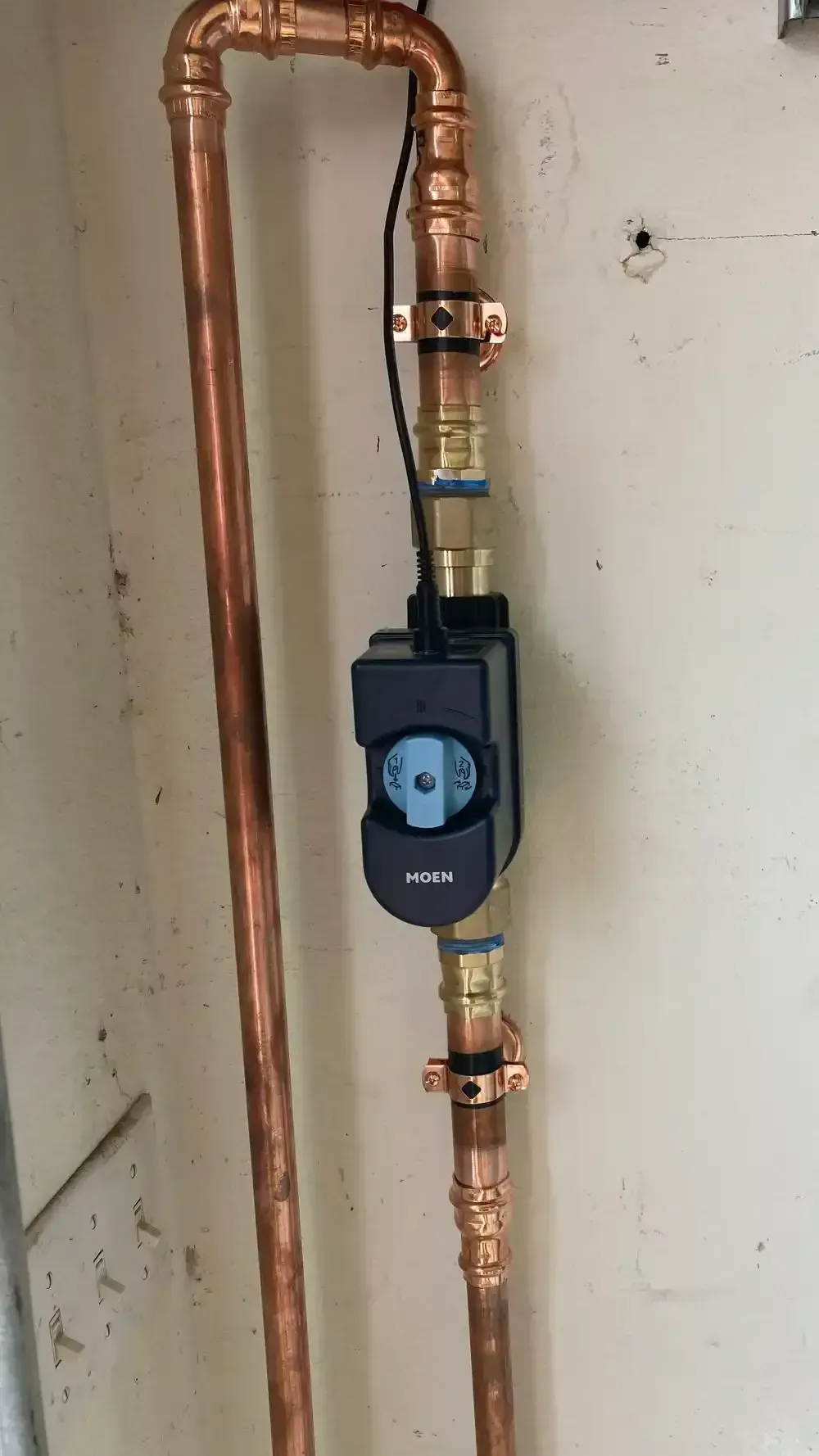 Copper pipes connected to a black control unit with a blue dial, mounted on a wall.