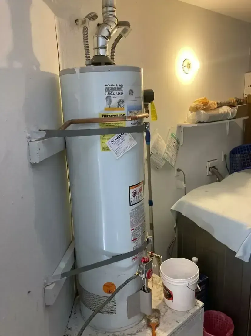 Water heater in a utility room, white tank, silver pipes, bucket below.