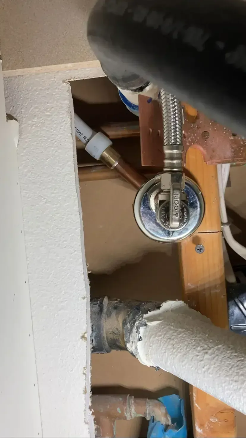 Plumbing under a sink: copper and PEX pipes, water valve, black hose, and wooden beams in a confined space.