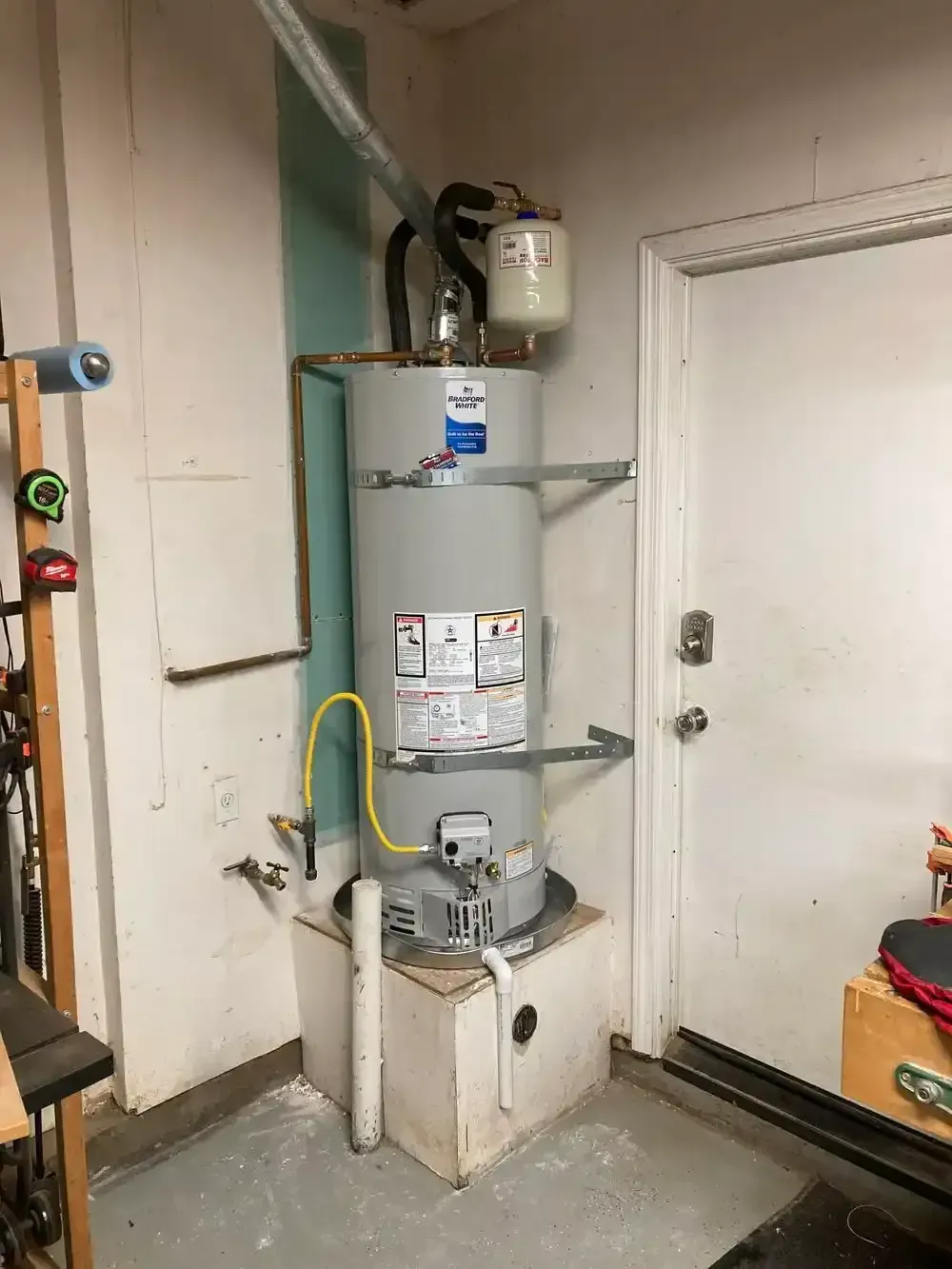Tall, gray water heater in a corner, with white door, shelves, and a yellow gas line.