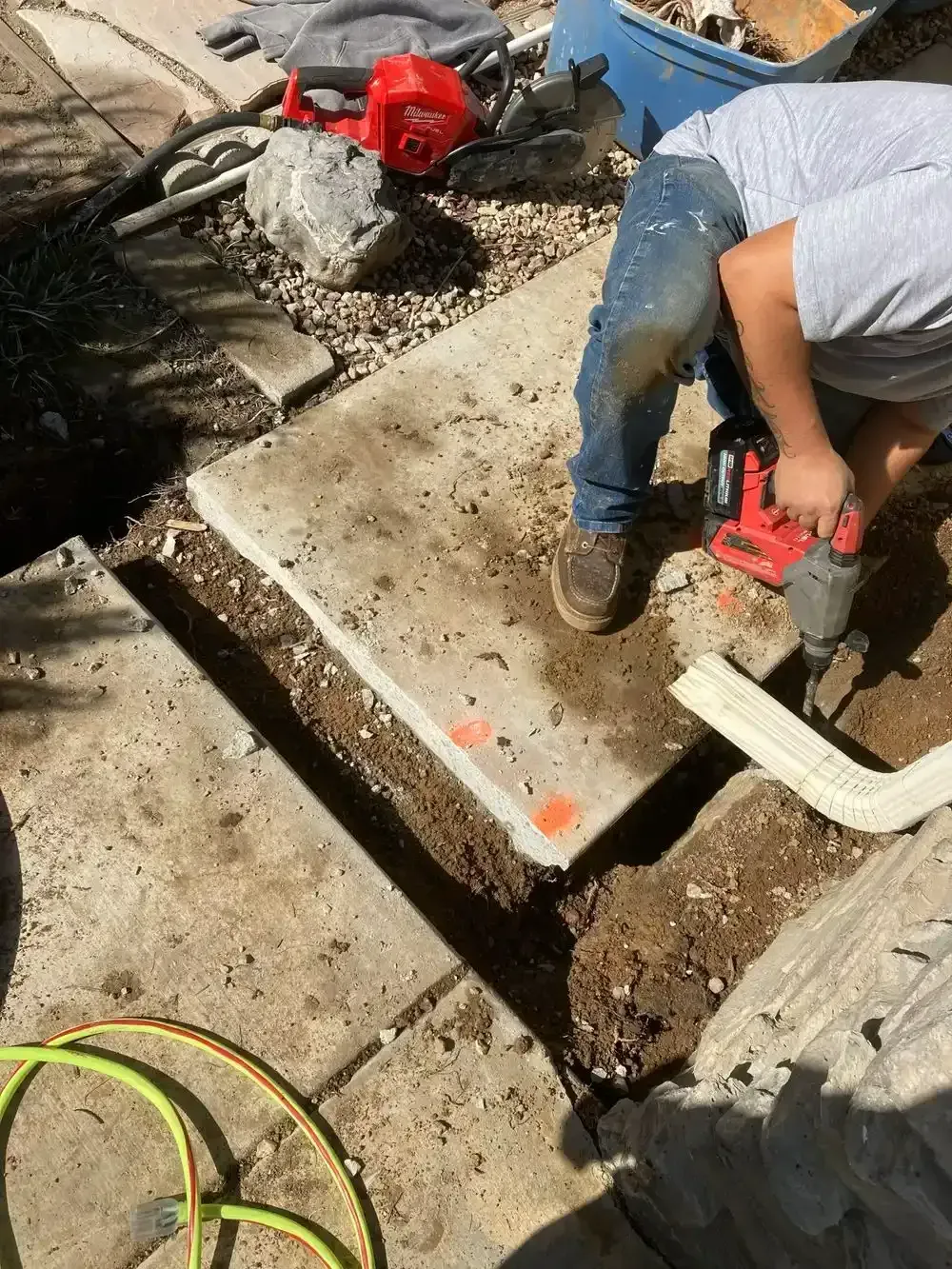 Person drilling into concrete walkway. A section of concrete is removed, exposing the soil.