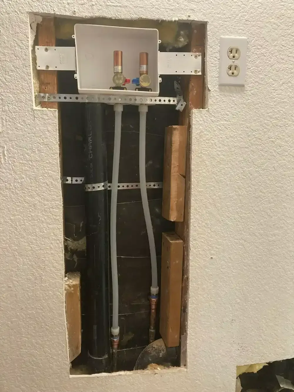 Plumbing for a washing machine in a wall cavity, including water lines, drain, and electrical outlet.