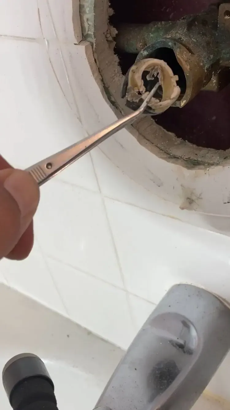A hand holding tweezers probing inside a damaged faucet fixture, likely for repair, near white tiles.