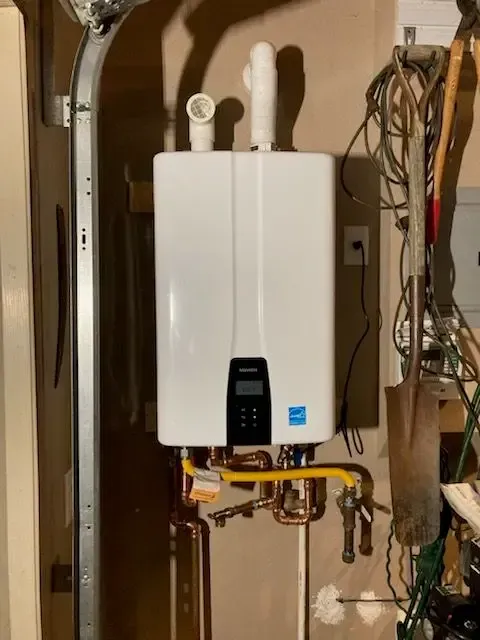 White tankless water heater mounted on a wall. Gas and water pipes connected. Garage setting.