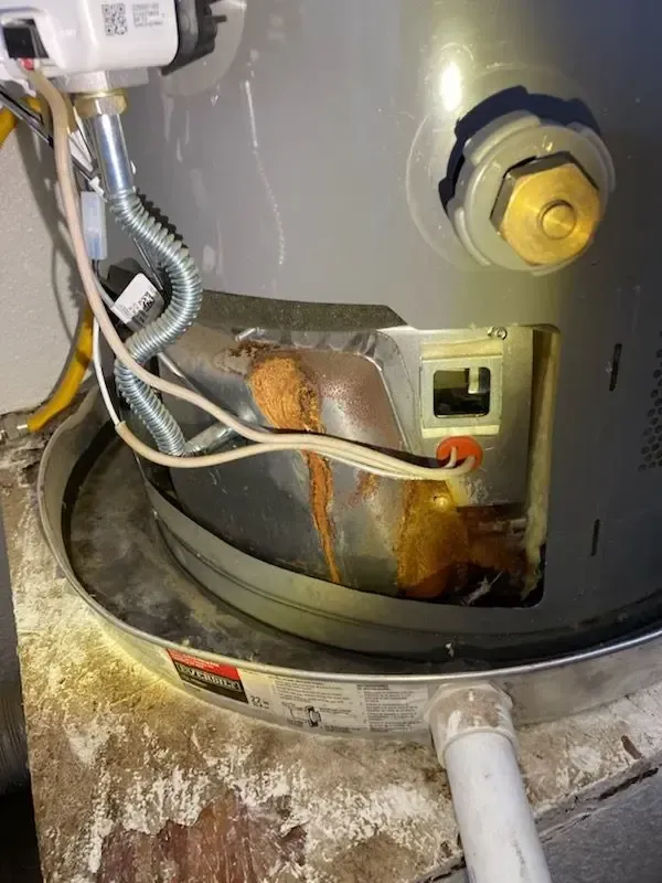 Corroded water heater showing rust and damage around the burner area; pipes and electrical components visible.
