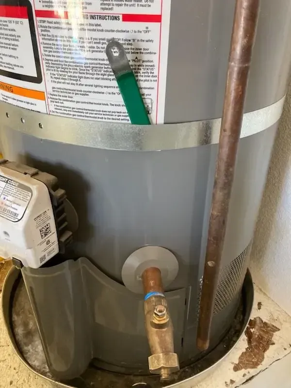 Gray water heater with copper pipes, a green valve handle, and a small amount of water on the floor.