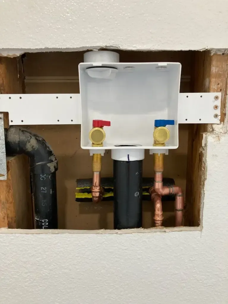 White laundry box with water supply valves and drain pipes in a wall opening.