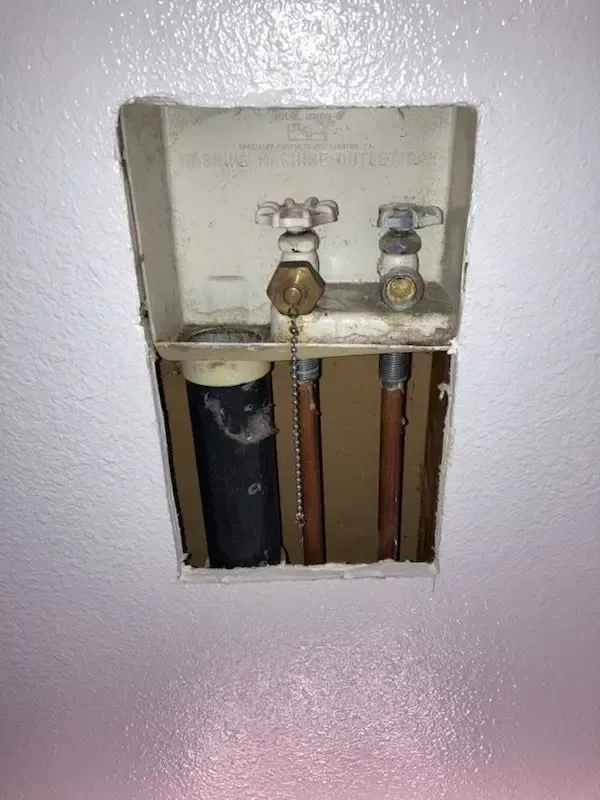 White wall opening with washing machine water valves, copper pipes, and drain pipe visible.