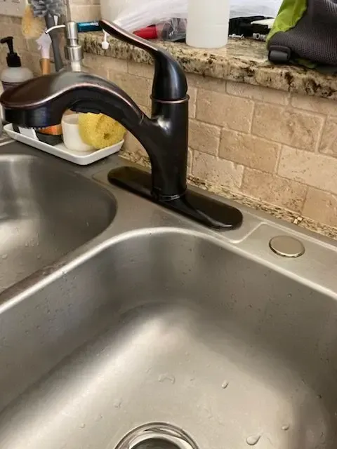 Bronze kitchen faucet mounted on stainless steel sink. Beige tile backsplash.