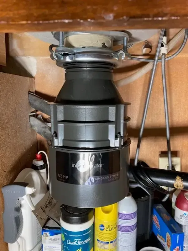 Garbage disposal unit mounted under a kitchen sink, surrounded by cleaning supplies and plumbing.