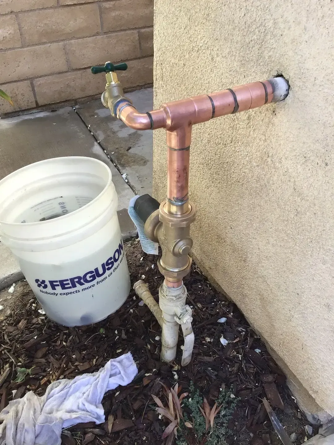 Copper pipes and outdoor spigot attached to a tan stucco wall next to a white bucket in dirt.